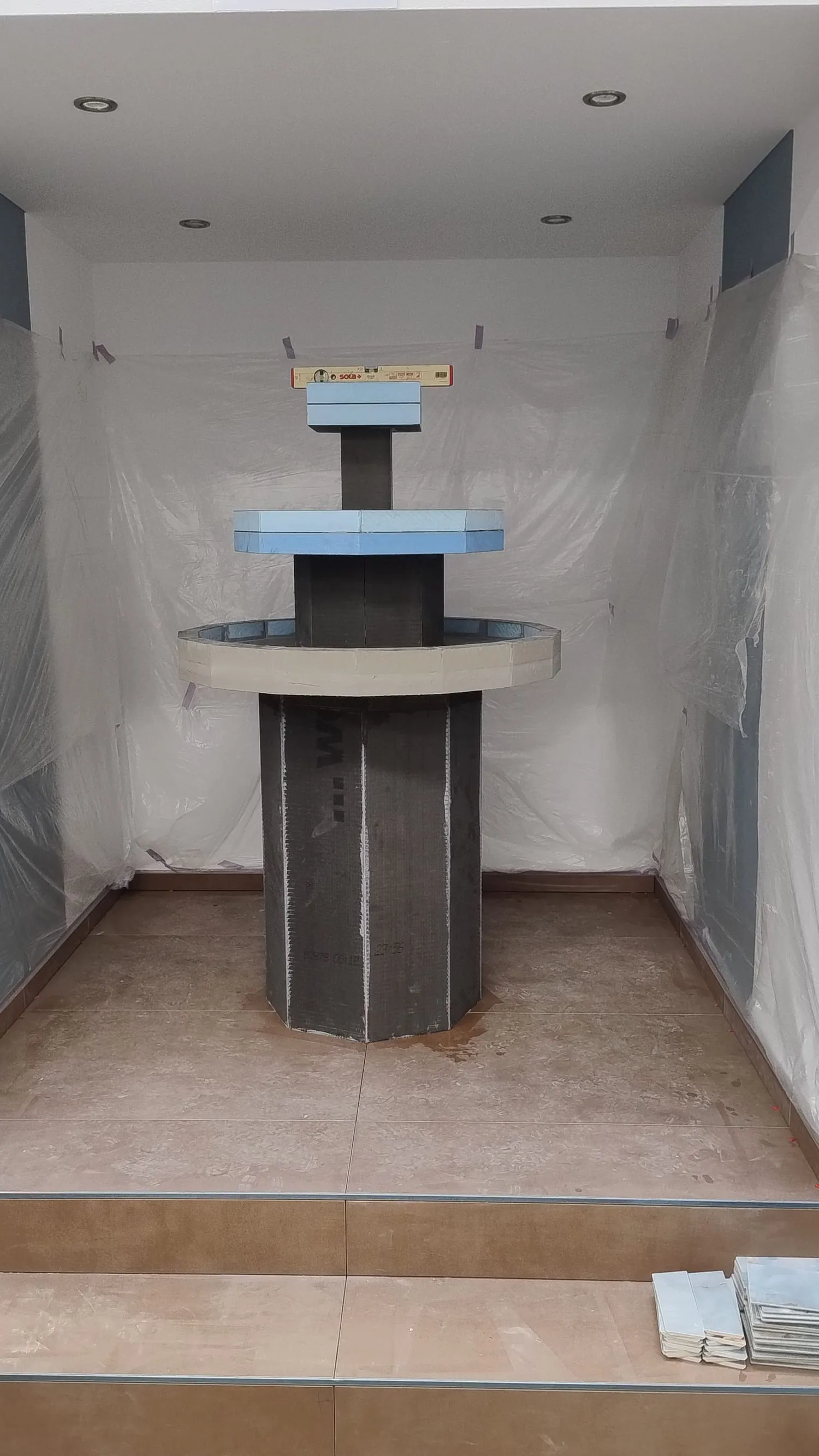Der Ausstellungsraum (Prüfungskoje) im Rohbau: Wände mit Folie abgedeckt, Trockenbaudecke mit Einbaustrahlern, zwei geflieste Stufen, davor der noch unverkleidete Brunnen