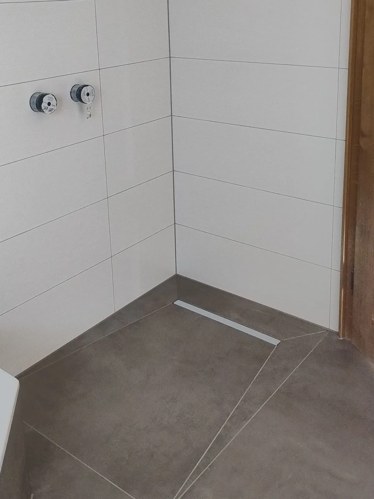 Bodengleiche Dusche mit weißen Wandfliesen, Armaturanschlüssen und Duschrinne im Boden